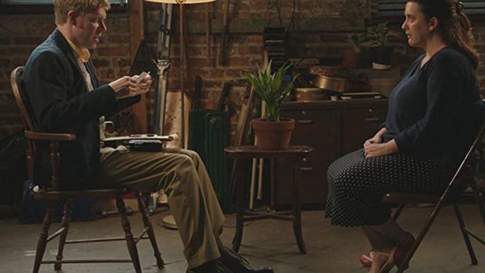 A man and a woman sit facing each other in a room with red brick walls.
