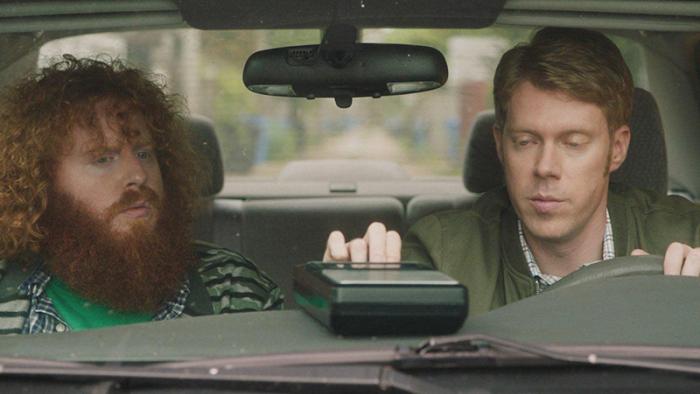 Two men sit in a car. The man in the passenger seat has long, curly red hair and a big red big. The man in the driver seat has short red hair and has his right hand on a cassette player that is sitting on the dashboard.