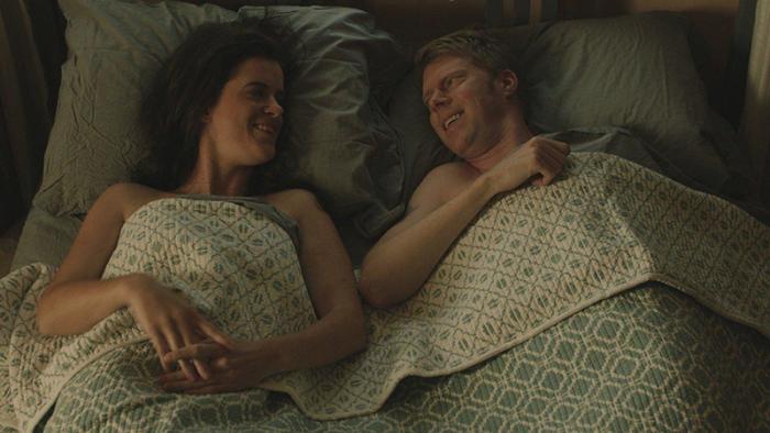 A woman and man are pictured laying in bed, under a blue and white patterned blanket, smiling at one another.