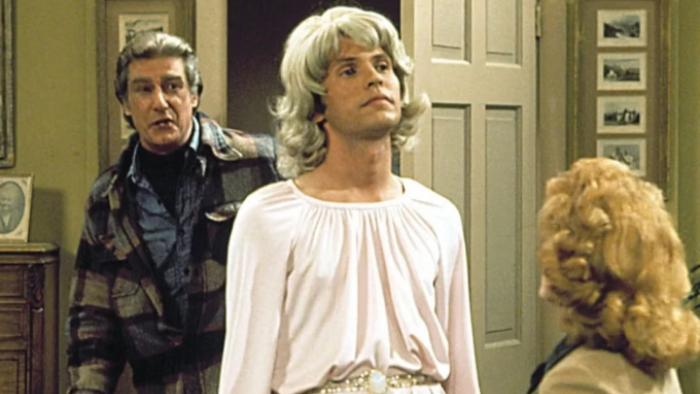 Soap: Jodie is center-screen, wearing a blonde wig and a pink dress; Burt stands behind him, looking disgusted, Mary's head is seen from behind