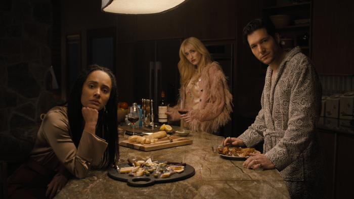 Karla Crome, Gus Birney and Jeff Wilbusch stand around a kitchen island. They have plates of food and glasses of wine in front.
