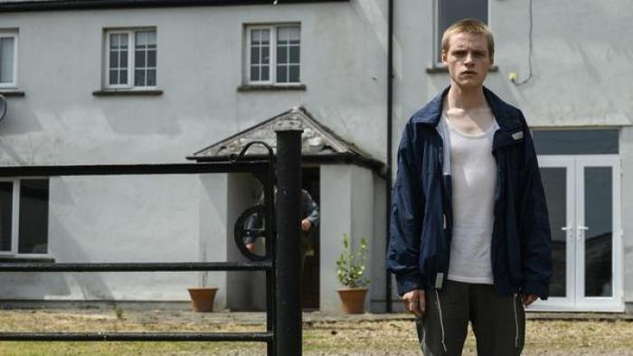 Somewhere Boy TV show screenshot 1: Lewis Gribben wears a white t shirt and blue jacket. He stands in front of a building.