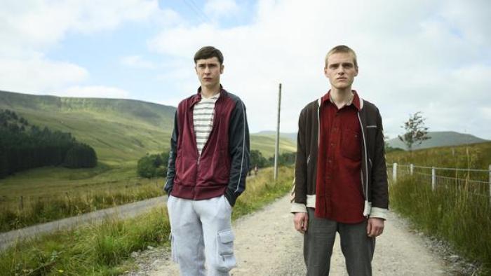Somewhere Boy TV show screenshot 2: Samuel Bottomley and Lewis Gribben wear red, standing in the middle of the road surrounded by a field.