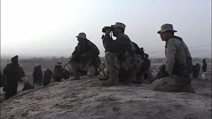 Armed soldiers sit on the ground outside on a dirt hill. One soldier holds up a pair of binoculars.