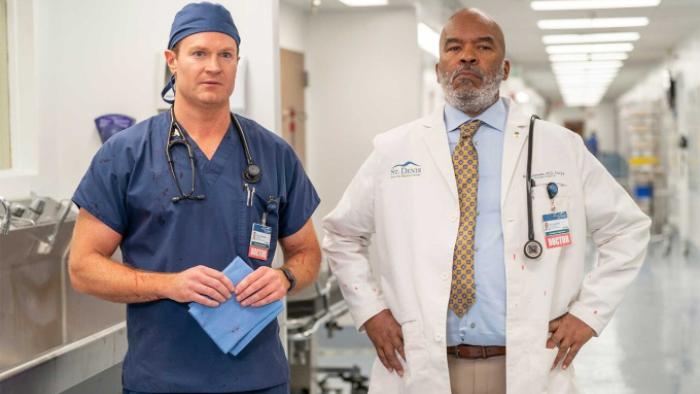 St. Denis Medical TV show screenshot 2: Bruce and Ron stand in a hospital corridor in scrubs and a white coat respectively, confronting a problem