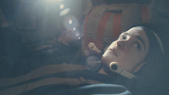 Alice Englert sits in spaceship cockpit. She wears a helmet, headphones, and a dark space suit.