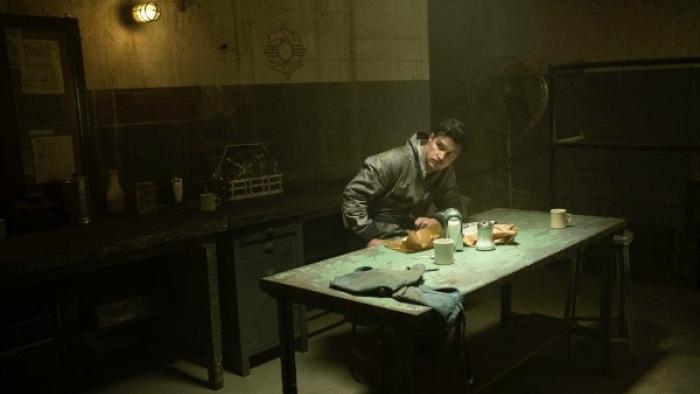 Strange Angel TV Series: A man in a dark prison-like room sits at a table hunched over, looking like he's upset with something he sees offscreen. 