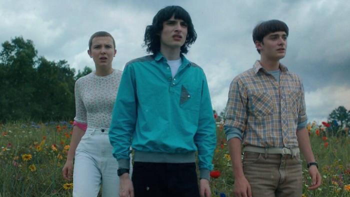 Stranger Things Season 5 TV show screenshot 1: Eleven, Mike and Noah stand on a grassy field with flowers and the blue sky behind them