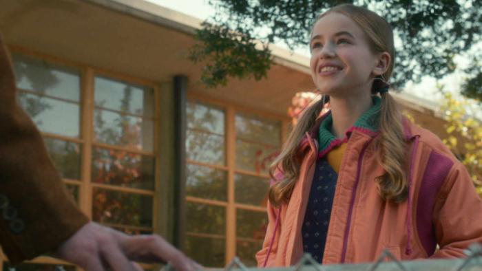 Stranger Things Season 5 screenshot 3: Holly smiles at a person off-camera; we see the hand of a young man in a brown suit from a person off-camera