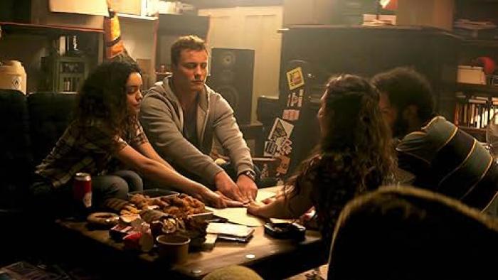 A group of four friends sit around a coffee table strewn with beer cans and ashtrays. Their hands rest atop a Ouija board.
