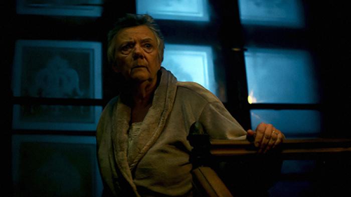 An older woman with grey hair stands at the top of a stairwell. She wears a velour bathrobe and has a concerned look on her face.