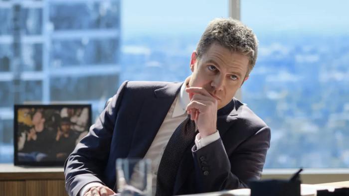 Stephen Amell sits at a desk, in the background are large windows showing a backdrop of L.A. Amell wears a dark suit.