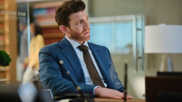 Bryan Greenberg sits at a desk. He wears a blue suit jacket, light blue shirt, and brown tie.