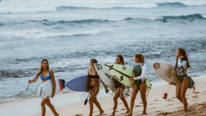 Surf Girls Hawai’i TV show screenshot 1: The women walk on the beach near water carrying surfboads.