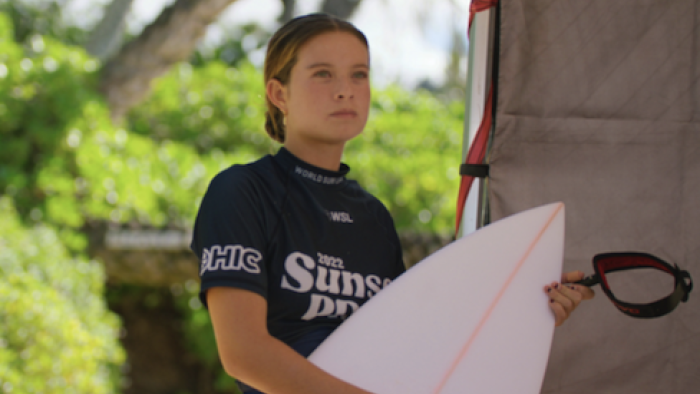 Surf Girls Hawai’i TV show screenshot-2: Pua in blue rash shirt holds white board. 