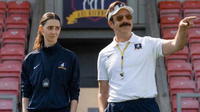Ted Lasso Season 4: Ted gestures at something on the football pitch as Alice stands beside him, looking irritated; both wear manager uniforms