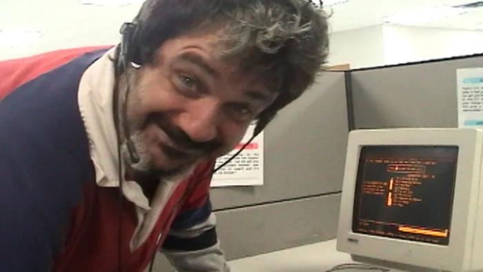 Telemarketers TV show screenshot 2: Pat Pespas leans down in front of a 1990s personal computer. He wears a phone headset and wears a striped shirt. He's smiling and appears as if he may be high on drugs. 