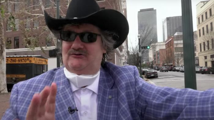 Telemarketers TV show screenshot 3: A man in a white shirt, gray suit jacket, and black cowboy hat is pictured in front of a city skyline. He holds up one hand as if to forestall someone from talking. 
