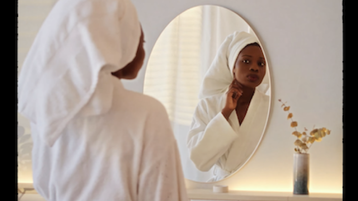 The Black Beauty Effect: A Black woman wrapped in white towel and robe examines face in mirror. 