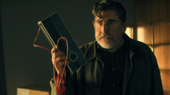 The Boroughs TV show screenshot 1: Sam stands in a darkened living room, holding a machine with a dangling plug with an angry expression on his face