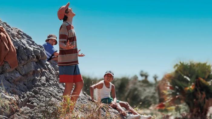 The Chosen One TV show screenshot 2: Against a desert background, Jodie, Magda, Wagner, Tuka and Hipólito are lounging on rocks. Jodie is standing and screeching something; his mouth is wide open. All are in casual summer clothing: shorts, t-shirts. 