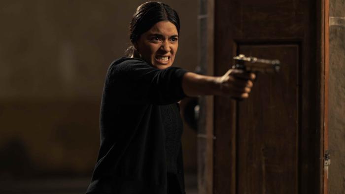 The Dead Girls TV show screenshot 1: Paulina Gaitán stands in the middle of open doors. She aims a handgun. She's dressed in black. 