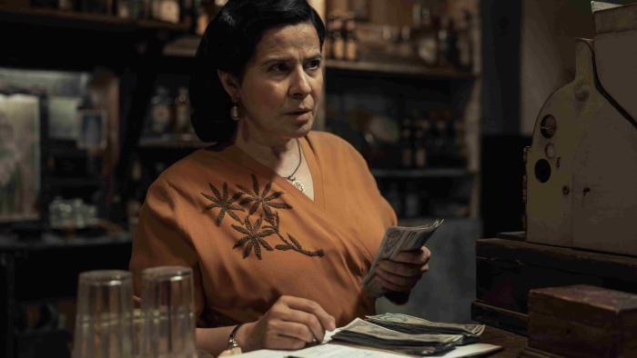 The Dead Girls TV show screenshot 2: Arcelia Ramírez holds money at a register. She wears a dark orange blouse with a brown floral on one side.