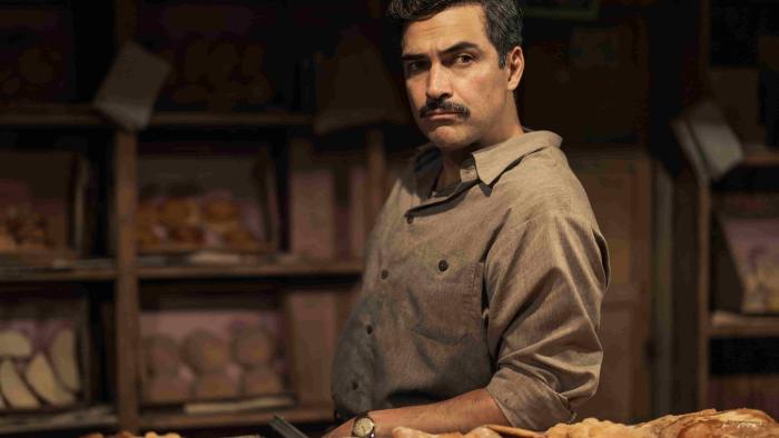 The Dead Girls TV show screenshot 3: Alfonso Herrera stands behind trays full of pastries. He wears a tan button-up shirt and a watch.