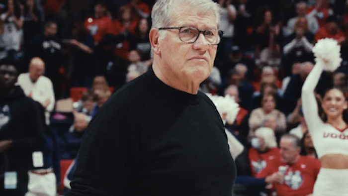 The Dynasty: UConn Huskies TV show screenshot 1: Coach Geno Auriemma in a black sweater stands on a basketball court, surrounded by cheering fans.