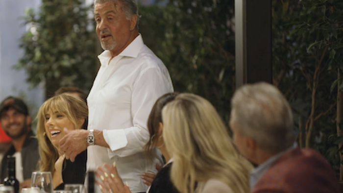 The Family Stallone screenshot 2: Sylvestor Stallone in white shirt stands and talks to table of dinner guests. 