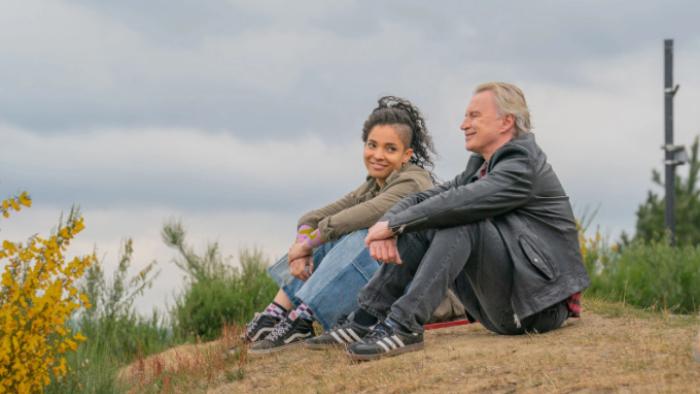 The Full Monty TV show: Destiny and Gaz sit on a hillside together, seemingly sharing a joke. Each wears jeans, sneakers, and a jacket. 