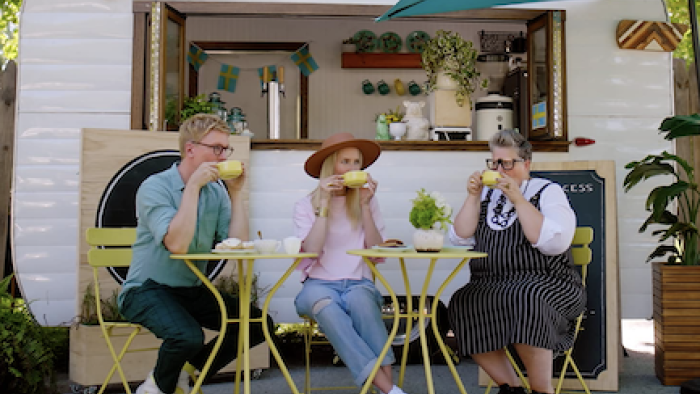 The Gentle Art of Swedish Death Cleaning screenshot 2: Johan, Kat, and Ella sip yellow coffee cups on yellow chairs in front of Frika stand.