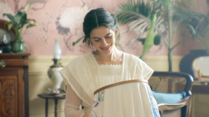 The House of the Spirits screenshot 1: Rosa smiling while embroidering in a vintage room with soft pink decor.