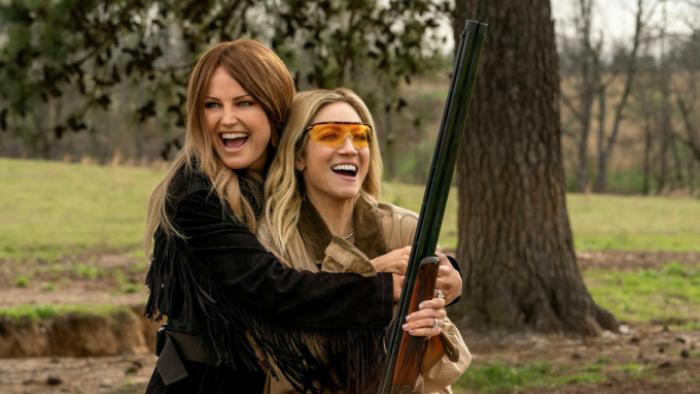 The Hunting Wives TV show screenshot 1: Margo holds Sophie in her arms while Sophie points a rifle into the air; both are in a woodsy area