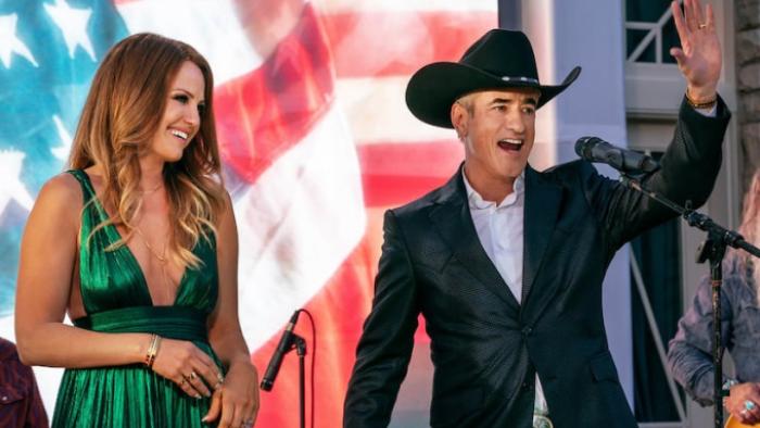 The Hunting Wives TV show screenshot 3: Margo and Jed are onstage in front of an American flag background; Margo smiles as Jed waves