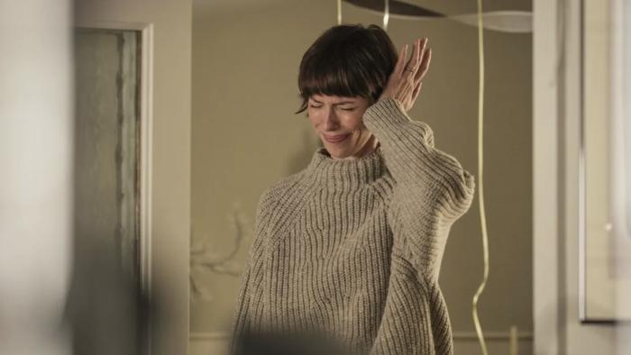 The Listeners TV show screenshot 3: Rebecca Hall holds one hand against her head, grimacing. She wears A chunky knit sweater.