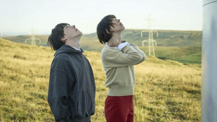 The Listeners TV show screenshot 2: Ollie West and Rebecca Hall stand in a field. West wears a dark hoodie; Hall wears a yellow sweater and red pants.