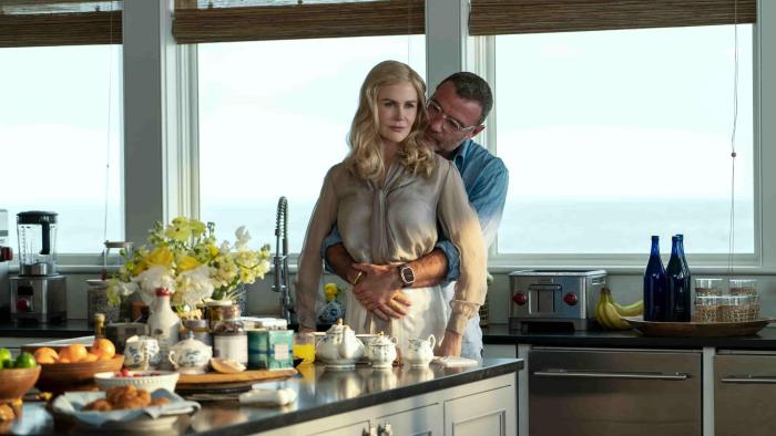 Liev Schreiber hugs Nicole Kidman while kissing her neck. They stand in a luxurious kitchen and the ocean in the background.