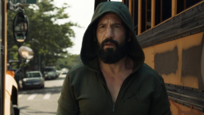 The Punisher: One Last Kill TV show screenshot 3: Jon Bernthal walks in between two yellow school buses. He wears a green zip up hoodie.
