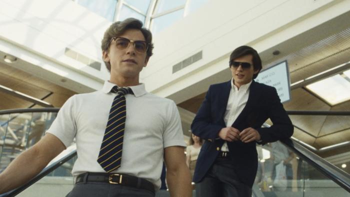 Igby Rigney and Homer Gere stand on a escalator in a mall. They both wear dark blue and white school uniforms, and sunglasses.