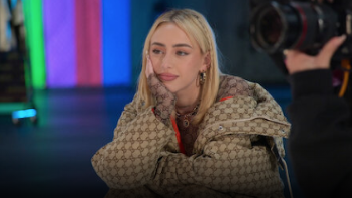 The Signing TV show: Nicki Nicole with brown jackets sits with her hand in her face