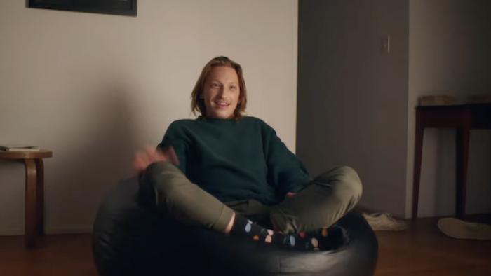 A blonde man with chin-length hair and colorful patterned socks sits cross-legged on a beanbag chair, seemingly mid-conversation.