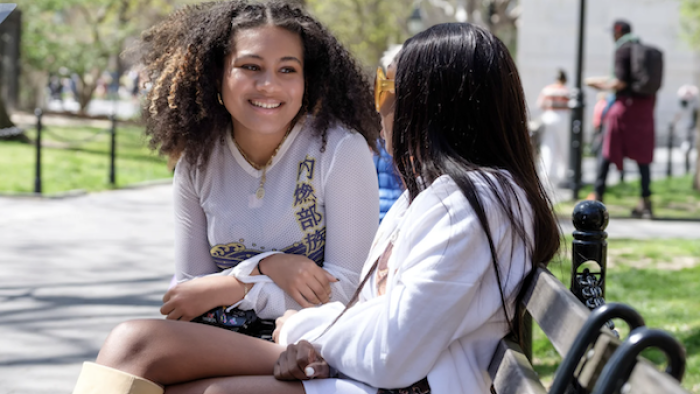 Two young women sit on a park bench. The woman at left has voluminous curly hair and is smiling at the women next to her, whose face we cannot see.