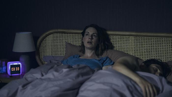 The Devil's Hour TV show screenshot 1: A woman sits awake in bed with a startled expression on her face. A man lays asleep next to her. The bedside cl