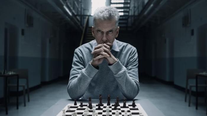 The Devil's Hour TV show screenshot 4: A man in a grey shirt is pictured sitting at a table with a chessboard in front of him. His hands are clasped a