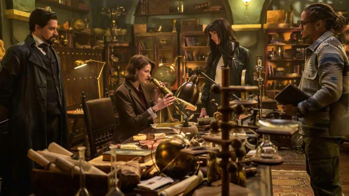 The Librarians The Next Chapter TV: In a warm-lit study room, Vikram, Charlie and Connor look to Lyra who sits with a telescope in a busy office desk.