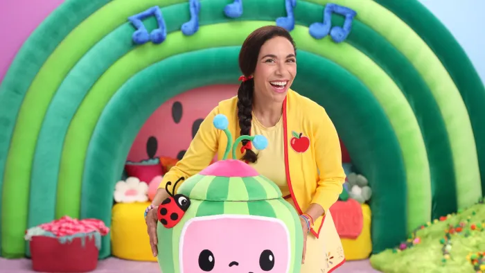The Melon Patch TV: Ms Appleberry stands behind the melon mystery box, in front of a melon rainbow, smiling at the camera.