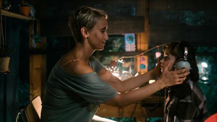 The Signal screenshot 3: Paula, wearing a loose grey t-shirt, smiles as she places a pair of headphones over Charlie's ears
