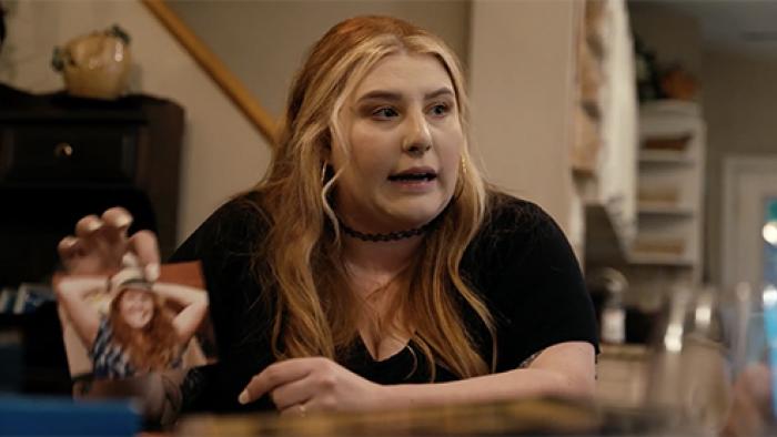 Thick Skin TV show screenshot 1: A white woman with long, red-and-blonde waby hair sits at a table, holding up a photo and speaking to someone off-camera.