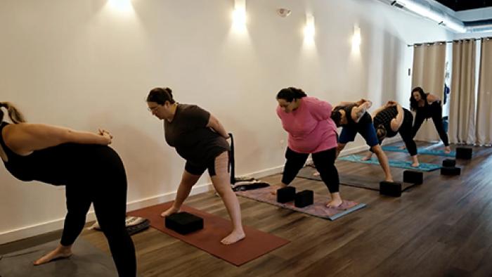 Thick Skin TV show screenshot 2: A row of six people in workout clothing stand over yoga mats with their hands clasped behind them, they are bent forward at the waist.